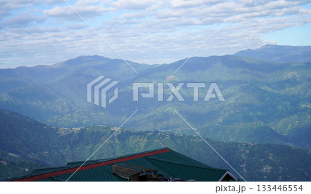 Green Rolling Mountain Range View with Distant Village Houses Under Cloudy Sky 133446554