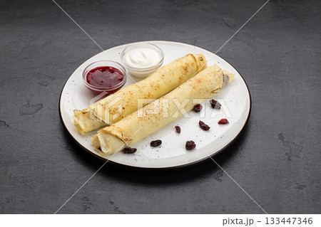 Rolled crepes with jam and sour cream on white plate, top view on dark background Rolled crepes with jam and sour cream on white plate, top view on dark background 133447346