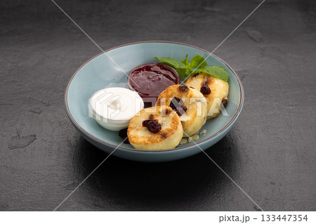 Syrniki pancakes with sour cream and berry jam in a blue bowl, traditional breakfast Syrniki pancakes with sour cream and berry jam in a blue bowl, traditional breakfast 133447354