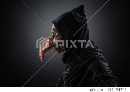 Creepy figure with long bird beak in hooded cloak over dark misty background 133454742