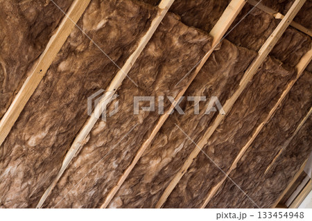 Brown glass wool in a wooden frame on a inclined wall near the wooden ceiling in a private house. Warming the house with fiberglass. Brown glass wool in a wooden frame on a inclined wall near the wooden ceiling in a private house. Warming the house with fiberglass. 133454948