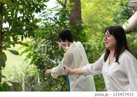 Two LGBTQ friends, one with long hair and the other with short hair, enjoy watering their garden in the morning. They share a joyful moment while caring for their plants in the lush outdoor space. Two LGBTQ friends, one with long hair and the other with short hair, enjoy watering their garden in the morning. They share a joyful moment while caring for their plants in the lush outdoor space. 133457413