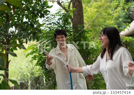 Two LGBTQ friends, one with long hair and the other with short hair, enjoy watering the garden together. They share a fun moment outdoors, taking care of their plants while laughing and bonding. Two LGBTQ friends, one with long hair and the other with short hair, enjoy watering the garden together. They share a fun moment outdoors, taking care of their plants while laughing and bonding. 133457414