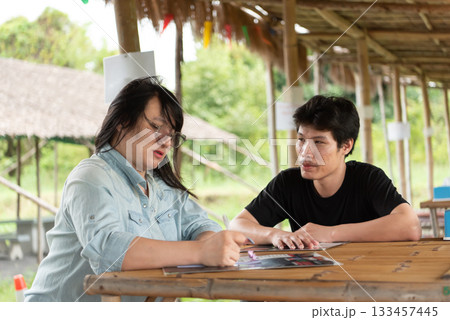 Two LGBTQ friends sitting at an Isaan Thai restaurant, enjoying their lunch together. One is writing notes while the other listens attentively, sharing a relaxed and enjoyable meal in cozy atmosphere. Two LGBTQ friends sitting at an Isaan Thai restaurant, enjoying their lunch together. One is writing notes while the other listens attentively, sharing a relaxed and enjoyable meal in cozy atmosphere. 133457445