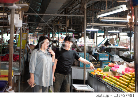 Two LGBTQ friends shopping for fresh vegetables at a local Thai market. One is pointing at the colorful fruits and vegetables, while the other is selecting produce, showing their lively interaction 133457481