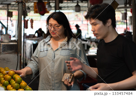 Two LGBTQ friends shopping for fresh fruits at a Thai market. One of them selects oranges while the other is holding a basket. The vibrant atmosphere of the busy market fills the scene. Two LGBTQ friends shopping for fresh fruits at a Thai market. One of them selects oranges while the other is holding a basket. The vibrant atmosphere of the busy market fills the scene. 133457484