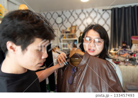 Two LGBTQ friends enjoy shopping for fashionable clothes at a boutique. One friend holds up a stylish leather jacket while the other discusses it, surrounded by trendy fashion items in a boutique. 133457493