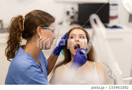 Girl is sitting in armchair during dental treatment at dentist appointment. Female doctor performs procedure of restoration and improvement of teeth, uses technological device Girl is sitting in armchair during dental treatment at dentist appointment. Female doctor performs procedure of restoration and improvement of teeth, uses technological device 133461892