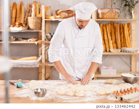 Professional baker stands at his work bench, kneading and shaping dough to make bread and baguettes Professional baker stands at his work bench, kneading and shaping dough to make bread and baguettes 133462442