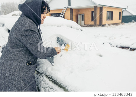 A young Hispanic woman with curly hair scrapes snow off a car in a winter setting. A house is visible in the background, covered in snow. 133463342