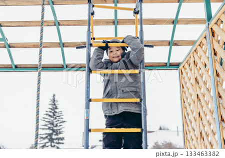 A young Asian boy climbs a yellow ladder in a snowy playground. Pine trees are visible in the background, creating a winter atmosphere, child safety on the street A young Asian boy climbs a yellow ladder in a snowy playground. Pine trees are visible in the background, creating a winter atmosphere, child safety on the street 133463382