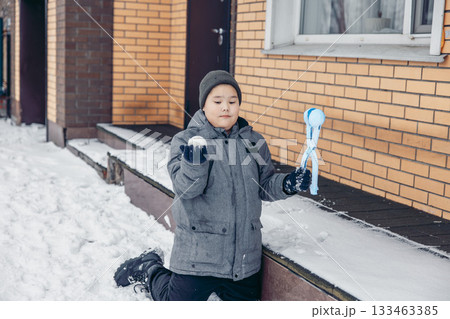 A young Asian boy kneels in the snow, holding a snowball in one hand and a snowball maker in the other. He wears a gray jacket and a beanie. 133463385