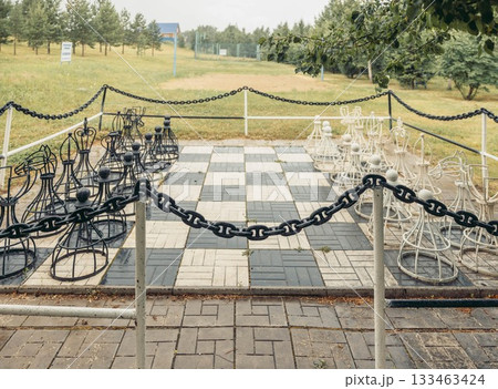 A large outdoor chessboard with decorative chairs as pieces. The board is surrounded by a chain fence. Green grass and trees are in the background. 133463424