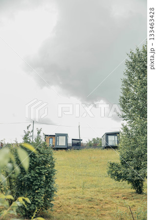 modern modular houses with large windows, on green hillside, grass, bushes under cloudy sky, contemporary architecture for eco living, countryside vacation, housing, tourism, nature 133463428