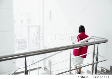 Unrecognizable woman in a red blazer walks down a modern staircase in a bright office environment. She carries a laptop and appears to be on a break. 133463454