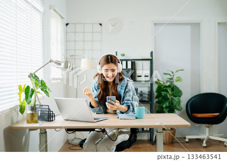 woman wearing headphones celebrating success while looking at smartphone in home office, symbolizing joy, digital achievement, and remote work lifestyle. 133463541