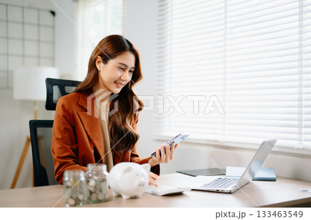 Asian businesswoman using smartphone and calculator for finance planning with piggy bank, coin jar, and laptop on desk Asian businesswoman using smartphone and calculator for finance planning with piggy bank, coin jar, and laptop on desk 133463549