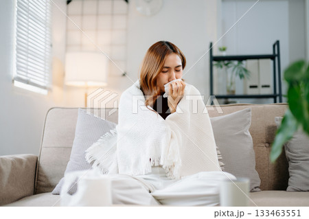 Young Asian woman suffering from flu symptoms, covered with a blanket on a sofa. Concept of illness, healthcare, and recovery. Stay indoors, stay healthy Young Asian woman suffering from flu symptoms, covered with a blanket on a sofa. Concept of illness, healthcare, and recovery. Stay indoors, stay healthy 133463551