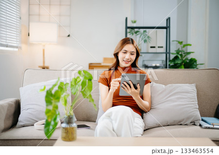 Happy Asian woman sitting on sofa and using digital tablet at home. Modern lifestyle, online learning, and technology concept for remote work and digital connection. Happy Asian woman sitting on sofa and using digital tablet at home. Modern lifestyle, online learning, and technology concept for remote work and digital connection. 133463554