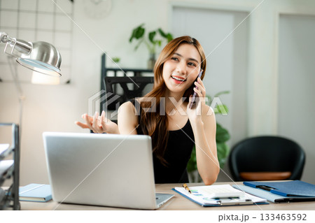Smiling businesswoman talking on phone while working on laptop in bright modern office, symbolizing success, communication 133463592