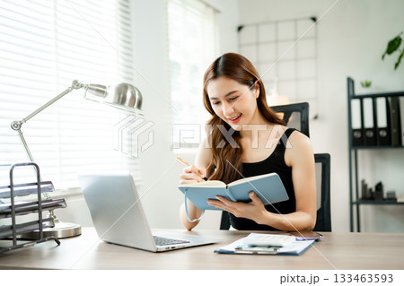 Smiling Asian woman working from home on laptop, writing notes, online learning or freelance business concept in home office. 133463593