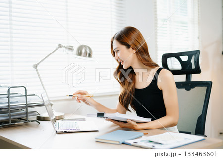 Smiling Asian woman standing and working with laptop in modern home office, concept of productivity, success, and female entrepreneurship. 133463601