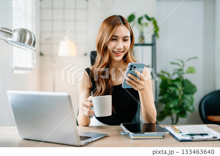 Happy businesswoman using smartphone and drinking coffee at laptop, symbolizing digital communication, modern lifestyle, and professional confidence. 133463640