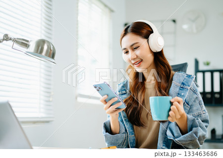 Smiling woman wearing headphones, enjoying coffee while working from home on laptop, symbolizing relaxation, creativity, and modern digital lifestyle. 133463686