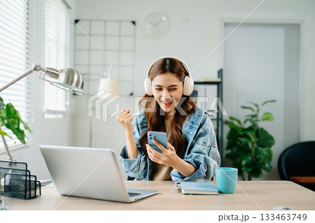 Excited woman wearing headphones celebrating success at laptop in bright home office Excited woman wearing headphones celebrating success at laptop in bright home office 133463749