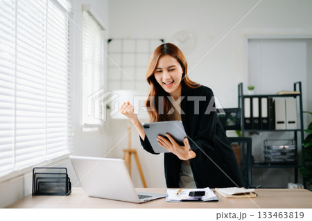 Asian businesswoman celebrating success at work, standing and cheering in modern office with laptop, showing joy, achievement, and victory in business and digital career. 133463819