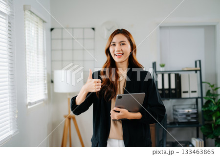 Confident Asian businesswoman smiling and giving thumbs up while holding tablet in modern office 133463865