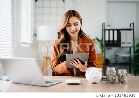 Young Asian businesswoman using tablet for finance and savings planning in modern office with piggy bank and coin jars, symbolizing money management, fintech, and success. Young Asian businesswoman using tablet for finance and savings planning in modern office with piggy bank and coin jars, symbolizing money management, fintech, and success. 133463939