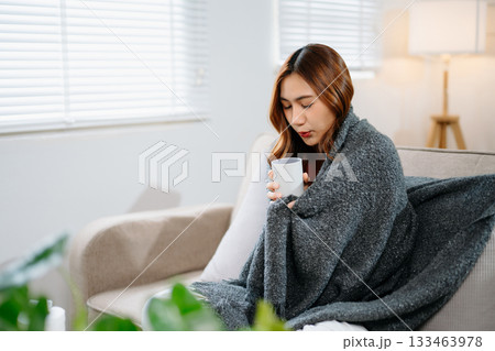 Young Asian woman suffering from flu symptoms, covered with a blanket on a sofa. Concept of illness, healthcare, and recovery. Stay indoors, stay healthy Young Asian woman suffering from flu symptoms, covered with a blanket on a sofa. Concept of illness, healthcare, and recovery. Stay indoors, stay healthy 133463978