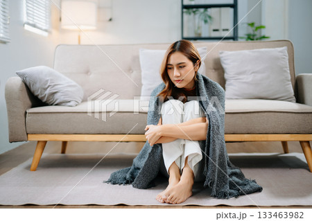 Sad Asian woman sitting on floor wrapped in blanket, holding head in distress. Concept of stress, anxiety, depression, burnout, and mental health problems. 133463982