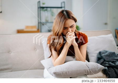 Sad Asian woman sitting on floor wrapped in blanket, holding head in distress. Concept of stress, anxiety, depression, burnout, and mental health problems. 133464050