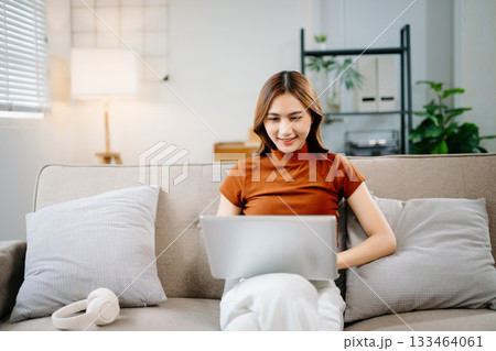 Cheerful Asian woman smiling while working on laptop at home. Modern remote work, online learning, and freelance lifestyle concept with confidence and creativity. 133464061