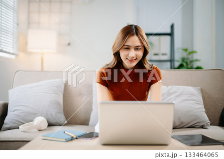 Cheerful Asian woman smiling while working on laptop at home. Modern remote work, online learning, and freelance lifestyle concept with confidence and creativity. 133464065