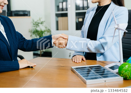 Two businesswomen shake hands in an eco office with wind and solar models, symbolizing green energy, clean tech, and sustainable business success. Two businesswomen shake hands in an eco office with wind and solar models, symbolizing green energy, clean tech, and sustainable business success. 133464582