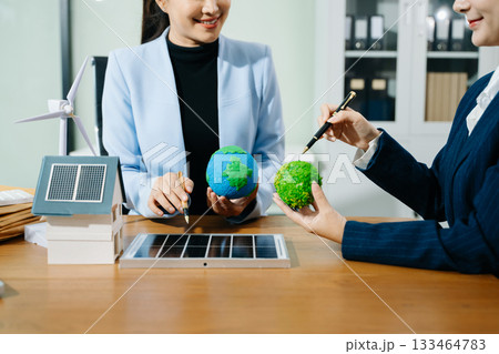 Two women analyze eco globe models, solar panels, and a smart home to plan renewable energy solutions for a greener, sustainable future. 133464783