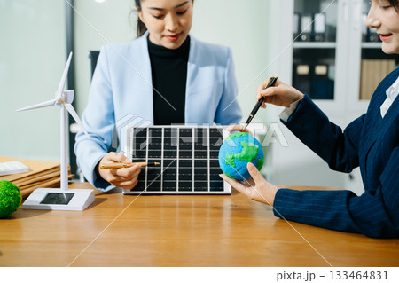 Two women analyze eco globe models, solar panels, and a smart home to plan renewable energy solutions for a greener, sustainable future. 133464831