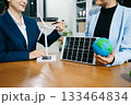 Two businesswomen shake hands in an eco office with wind and solar models, symbolizing green energy, clean tech, and sustainable business success. 133464834