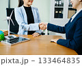 Two businesswomen shake hands in an eco office with wind and solar models, symbolizing green energy, clean tech, and sustainable business success. 133464835
