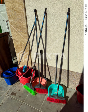 Colorful brooms and buckets resting against a wall in a sunlit corner of a bustling marketplace during a busy afternoon Colorful brooms and buckets resting against a wall in a sunlit corner of a bustling marketplace during a busy afternoon 133469346