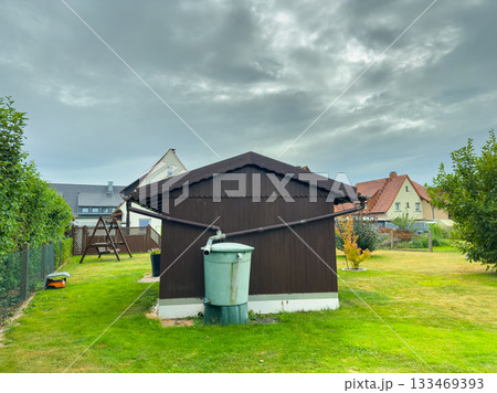 Charming garden scene featuring a brown wooden shed with a green water tank under a cloudy sky in a peaceful neighborhood 133469393