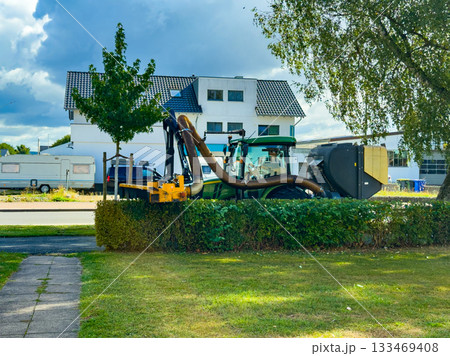 Busy morning with a large green machine cleaning up the street in a quiet neighborhood under a partly cloudy sky 133469408
