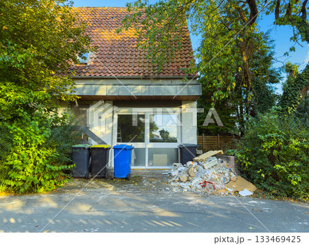 Abandoned house surrounded by greenery with debris and trash bins on a sunny afternoon Abandoned house surrounded by greenery with debris and trash bins on a sunny afternoon 133469425