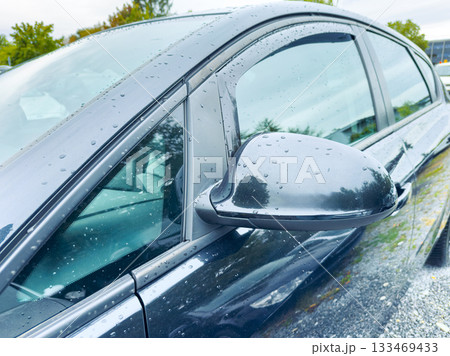 Shiny black car reflects light and raindrops during a cloudy day in a serene parking lot surrounded by lush greenery Shiny black car reflects light and raindrops during a cloudy day in a serene parking lot surrounded by lush greenery 133469433