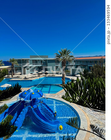 Bright blue waters flow from a fun slide into a sparkling pool surrounded by palm trees and elegant accommodations under a clear sky 133469594