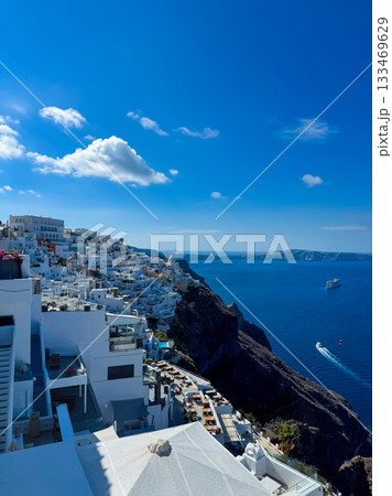 Beautiful coastal view of white buildings against the deep blue sea in Santorini with boats sailing in the distance during a bright sunny day 133469629