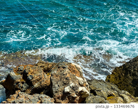 Waves crashing against rugged rocks at a vibrant coastal landscape under bright sunlight Waves crashing against rugged rocks at a vibrant coastal landscape under bright sunlight 133469676
